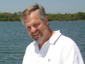 Head-and-shoulders shot of a man smiling in front of a sunny water scene