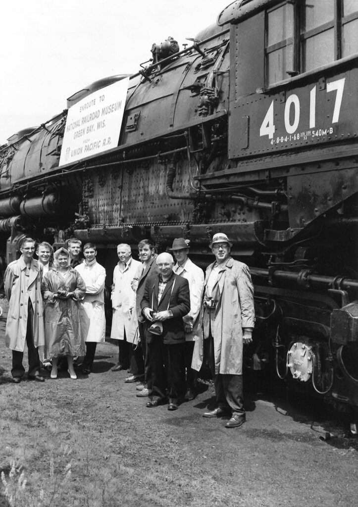 Black-and-white photo of people standing in front of steam engine.