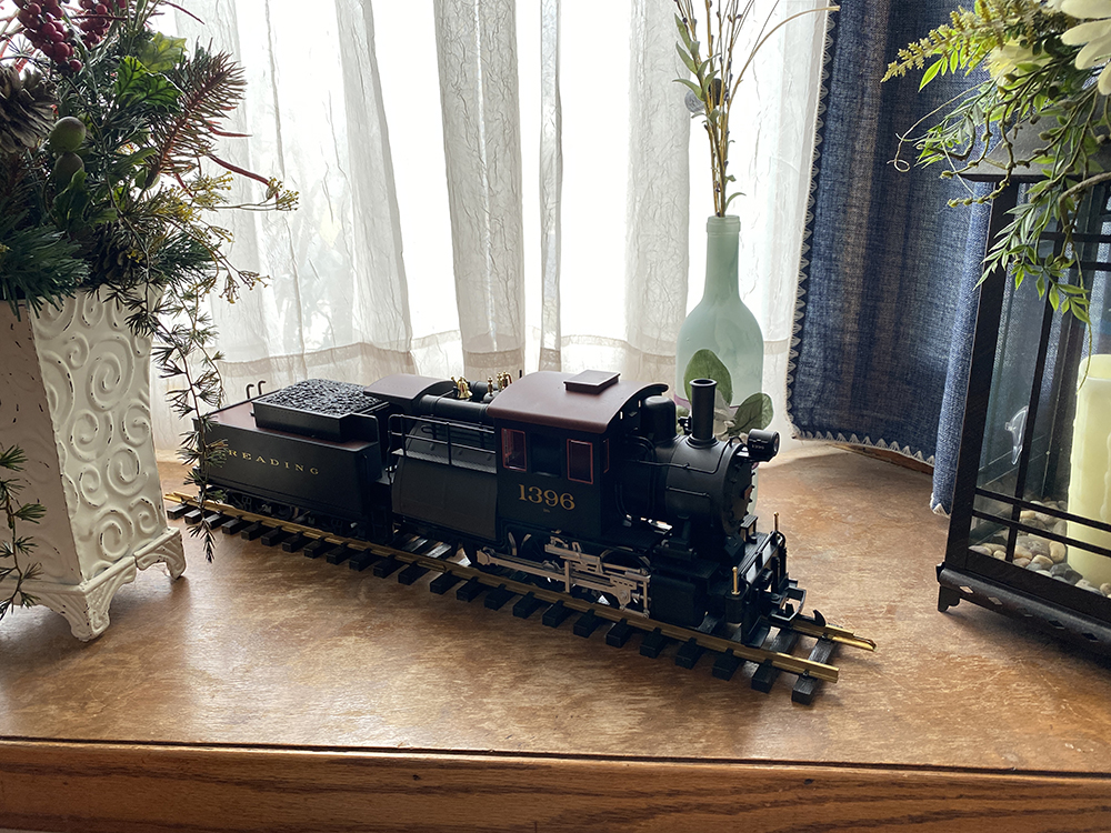 Black model steam locomotive on brass track resting on brown wood shelf and surrounded by light-colored vases filled with plants.