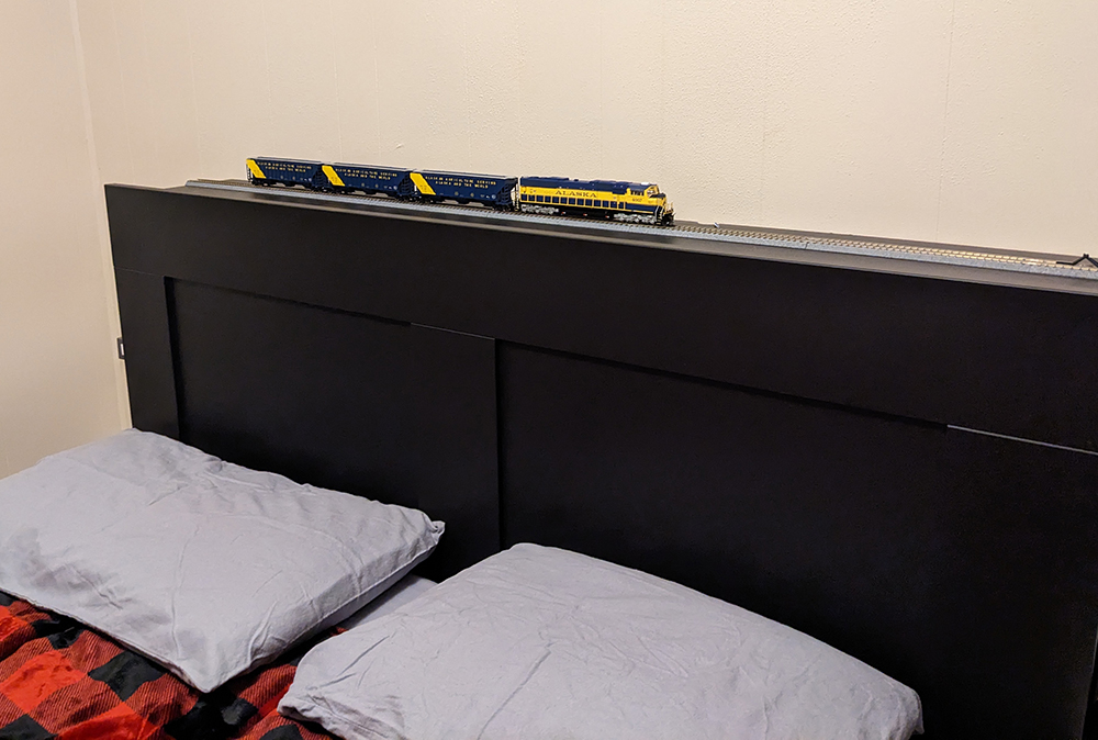 Gray model train track on black headboard of a bed with red pillows