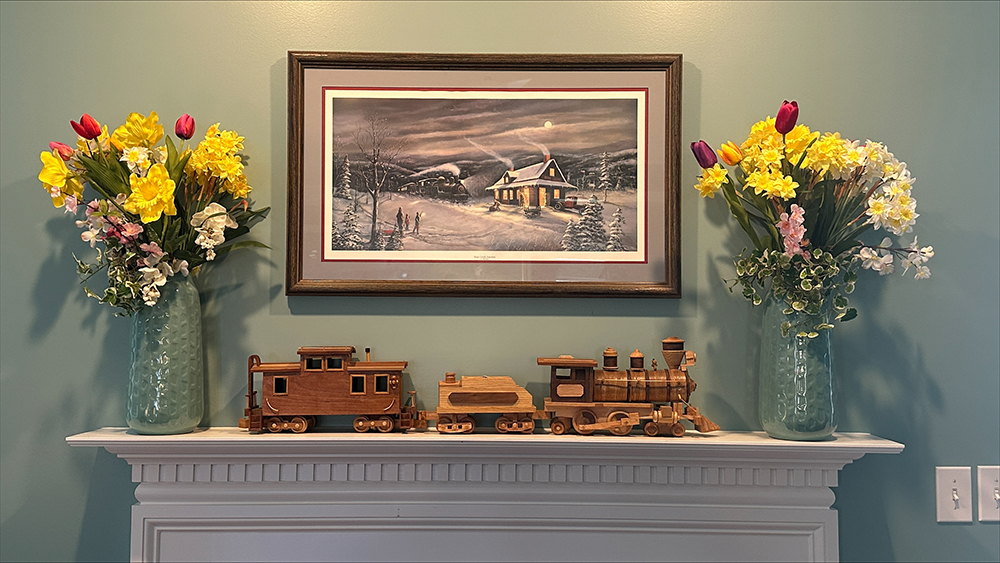 Wooden steam train with caboose displayed on a white fireplace mantle and surrounded by yellow flowers