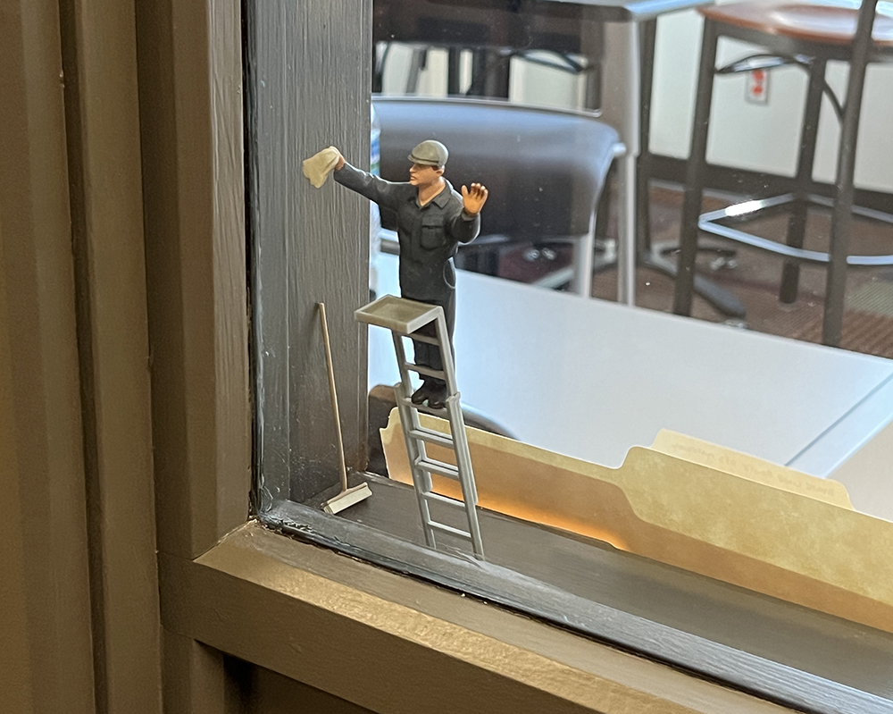 toy plastic man in blue overalls standing on ladder and attempting to wash a full-sized windowpane