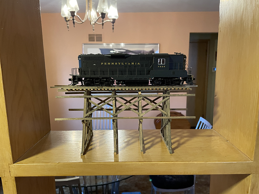 Black model train engine with gold lettering on small wood trestle on brown bookshelf