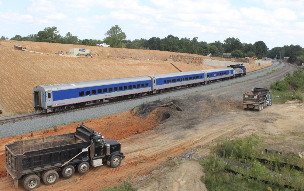 Passenger train passes freshly graded dirt and dump trucks