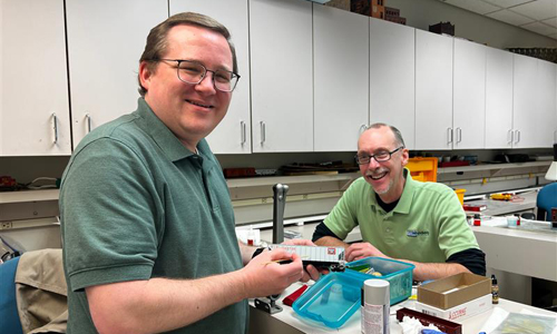Two men at a model railroading workbench smiling at a camera.