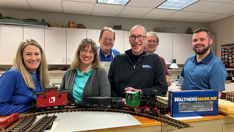 Several people smile into a camera at a model railroad layout.