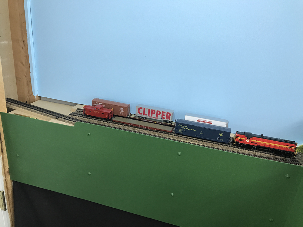 Model train track on a green shelf mounted to a blue wall supporting red, blue, silver, and brown railroad equipment