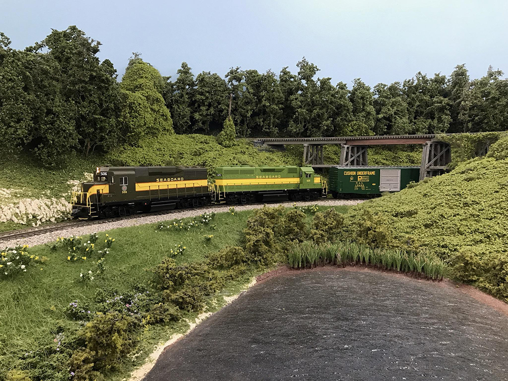 Model train set on a table with model lake in foreground surrounded by green grass and weeds, a green and yellow train on track with tan ballast behind it, and trees and a brown wooden railroad trestle in the distant background