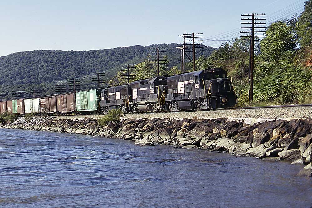 Three black-and-white diesel Penn Central locomotives on freight train along river
