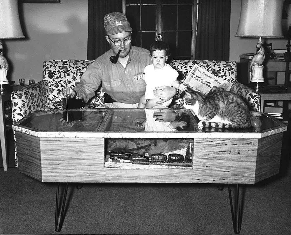Man with pipe holding baby and looking at table with cat on it