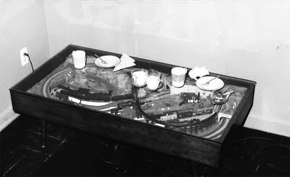 Black and white image showing table in corner of room with white cups, plates, and napkins on it.