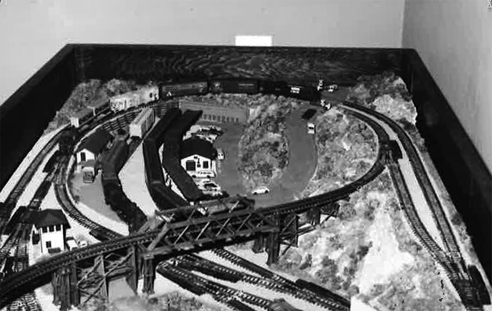 Black and white photo of curved model train track on table with landscape, miniature buildings, vehicles, and a wooden bridge