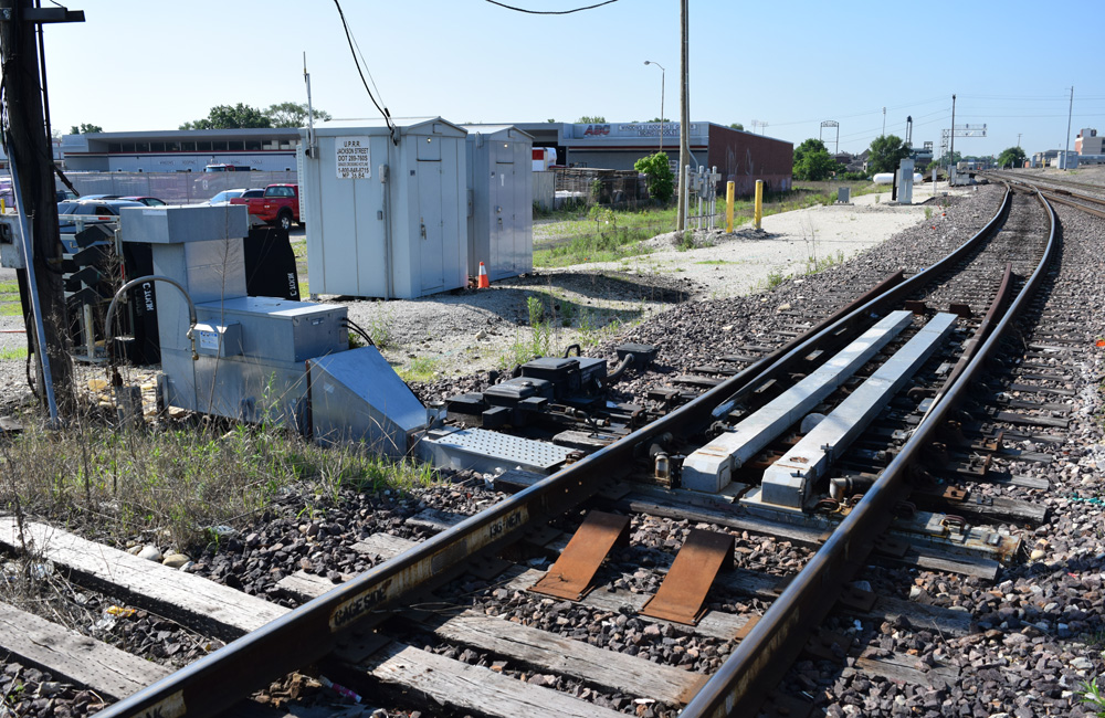 A boxy metallic device with a rectangular chimney stands next to a section of railroad track with two metallic ducts extending between the rails