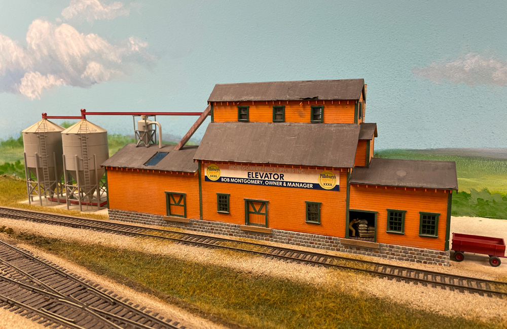 An orange, wood-sided feed mill with attached grain bins is seen on an HO scale train layout