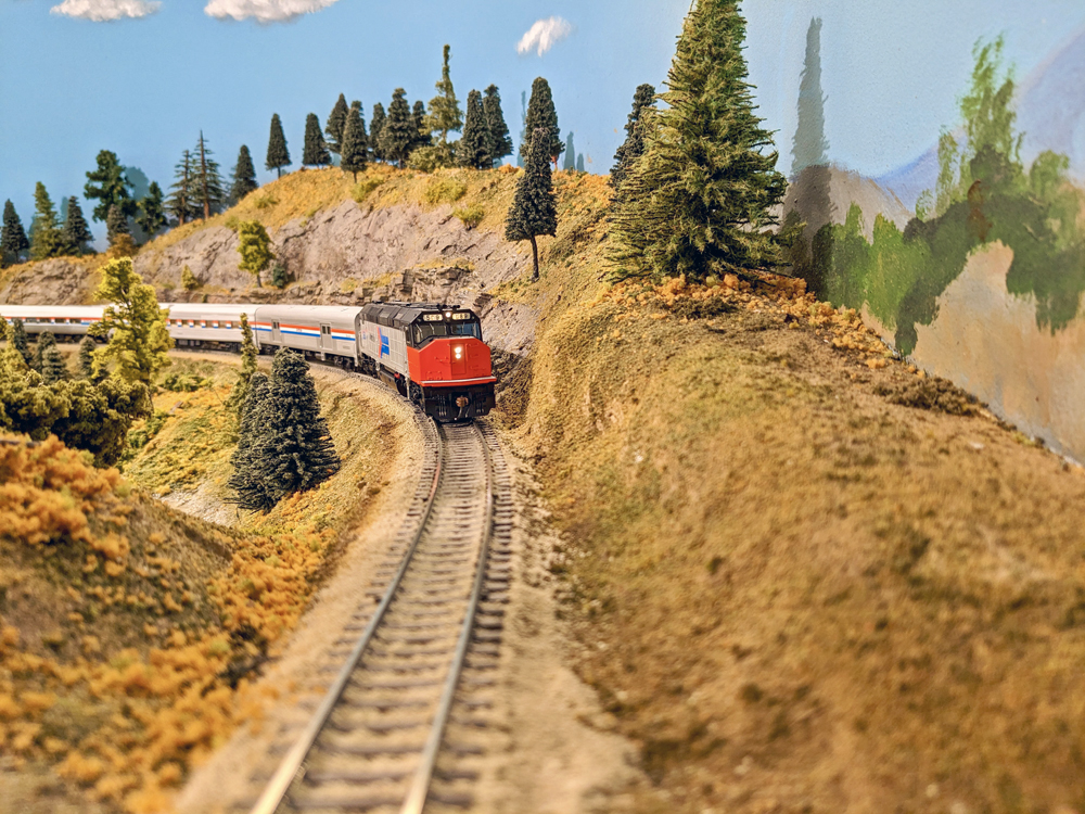 A model passenger train heads down the tracks, through a mountain cut.