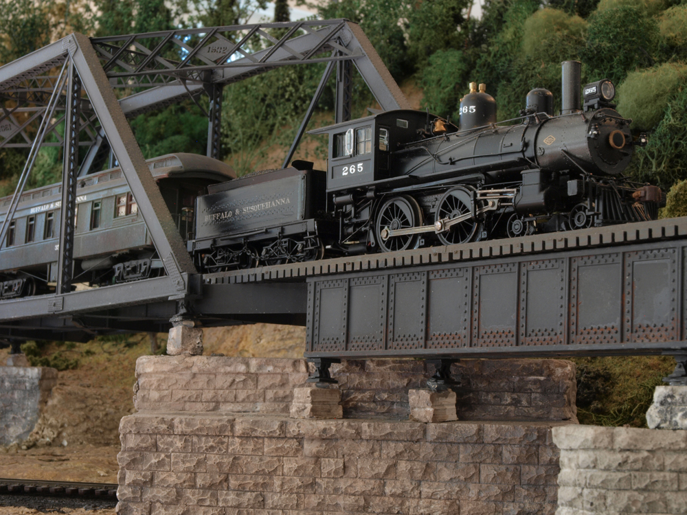 A model steam locomotive pulls a passenger car over a bridge.