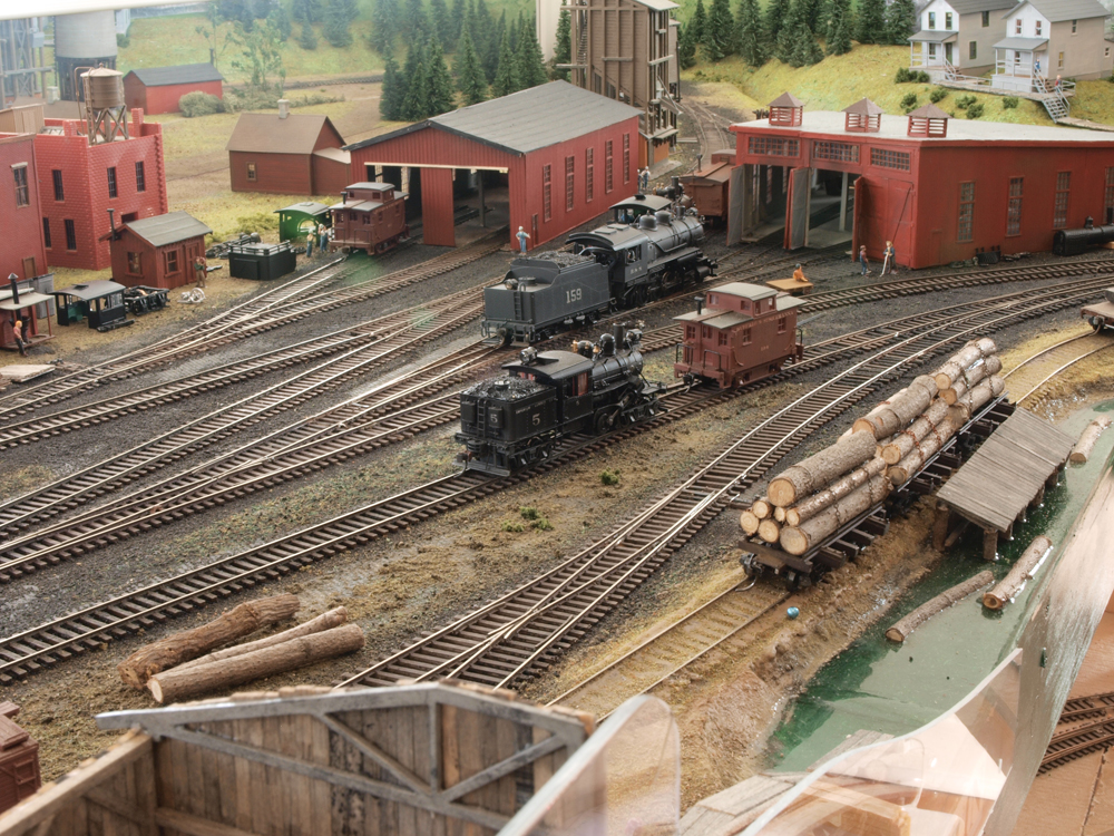 Model steam locomotives and a caboose sit in a yard with shop buildings in the background.