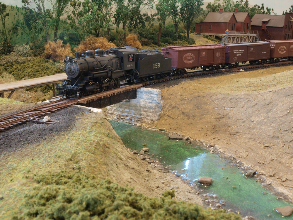 A model steam engine pulls boxcars over a small bridge.