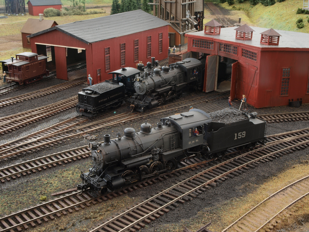 Model steam locomotives sit in a yard with shop buildings in the background.