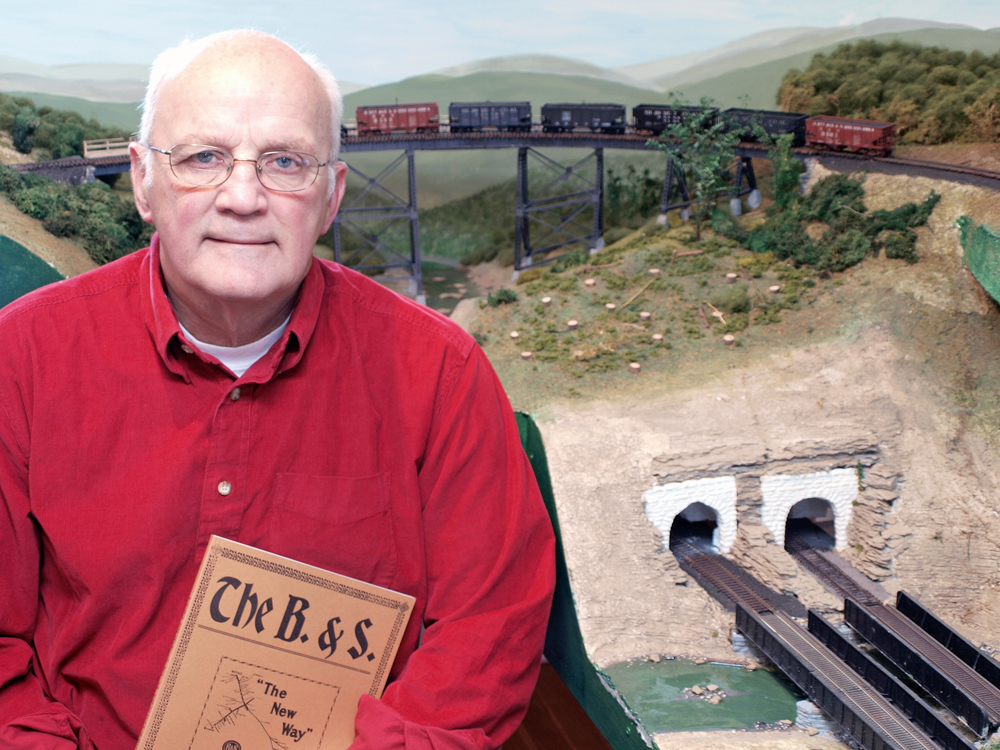 Photo of a man with a book in his hand in front of a model train layout.