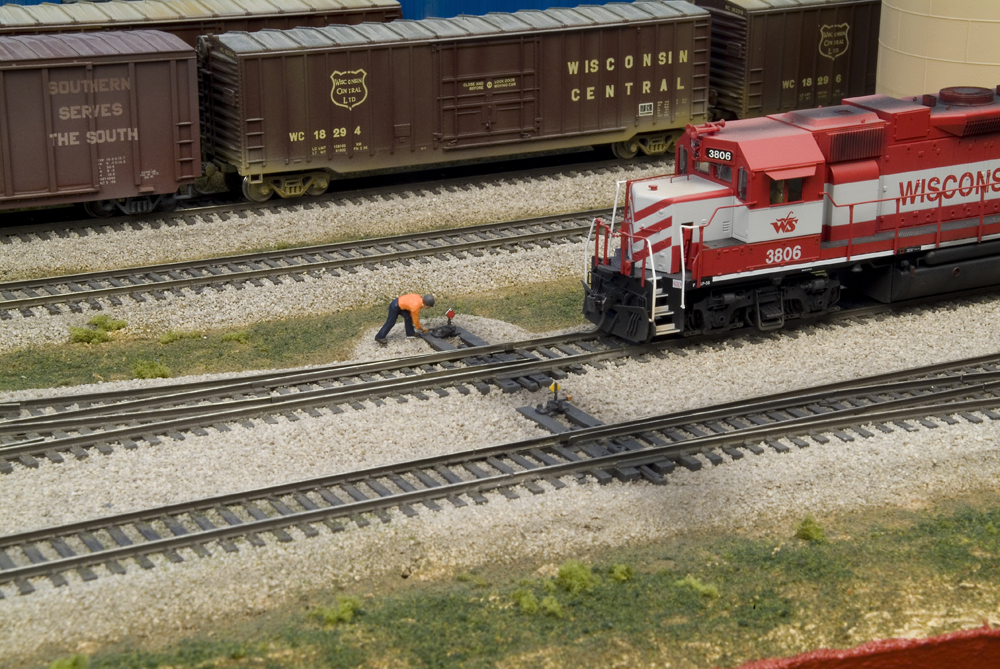 Color photo of HO scale figure by turnout with locomotive and boxcars in the frame. 