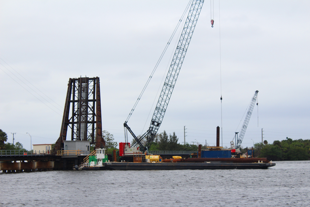 Cranes on barge in river next to railroad drawbridge.