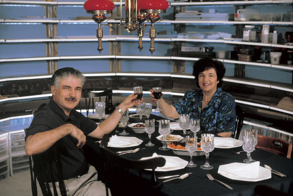 Model Train Décor Using Shelf Layouts: A man and woman seated at a table and holding wine glasses surrounded by white shelves in the background.