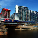 Passenger train with red and blue locomotive with body of water in foreground and high-rise apartments behind