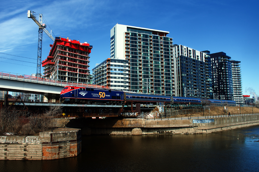 Passenger train with red and blue locomotive with body of water in foreground and high-rise apartments behind