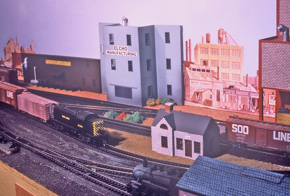 Built by Others: Bob Wundrock’s HO scale Gold Hill Central: Yellow, black, and brown toy trains in a town setting with gray and brown buildings
