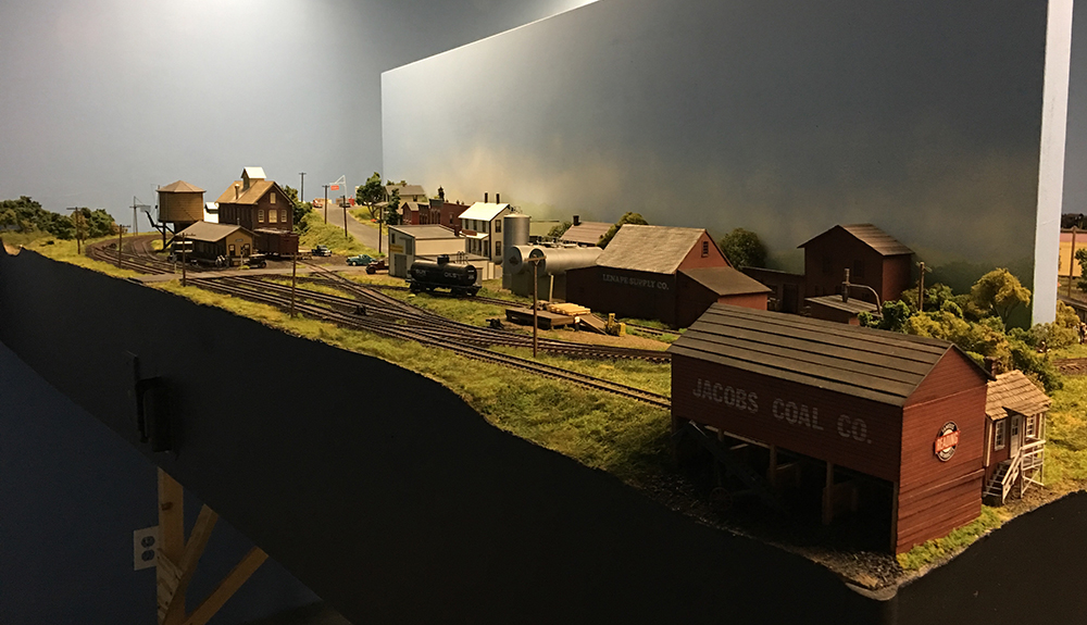 Merkiomen Valley Branch layout: Model train layout with green grass, brown track, tan and red buildings, and a blue sky