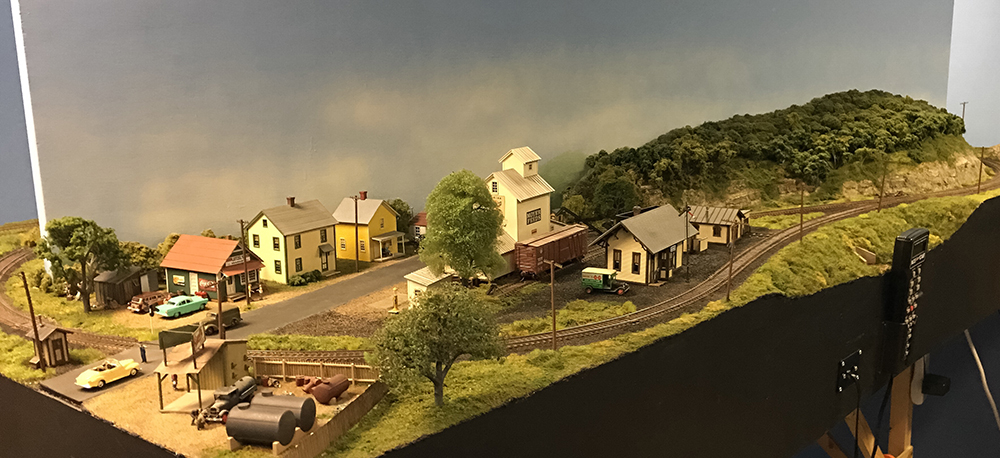 Merkiomen Valley Branch layout: Small model railroad town featuring yellow and tan houses, a white feed supply building, and a small tan and green depot.
