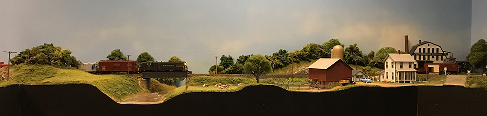 Model train set featuring green grass and trees, brown water, black bridge, red barn and white farmhouse.