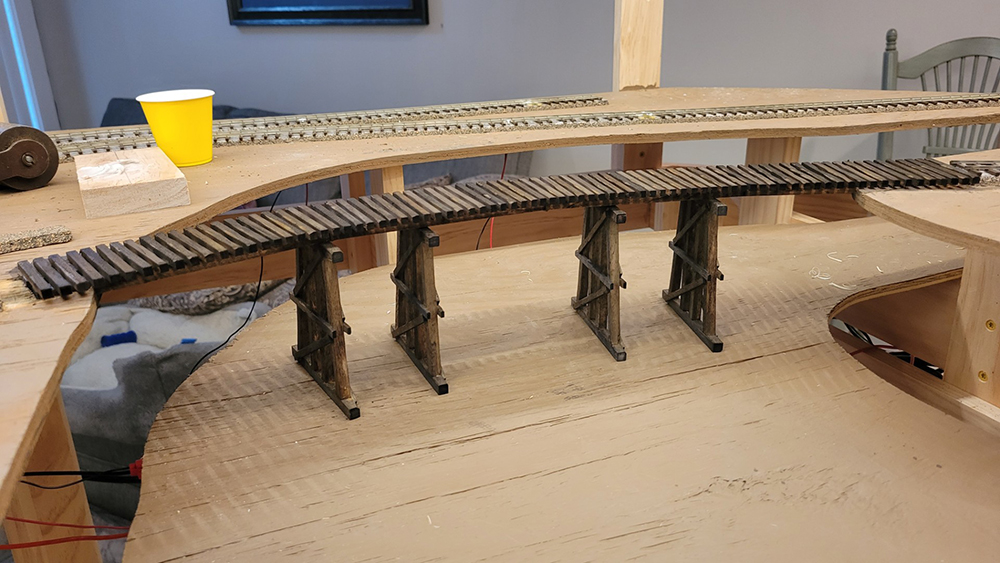 An unfinished wood model railroad curved trestle without rails sitting on tan plywood surface