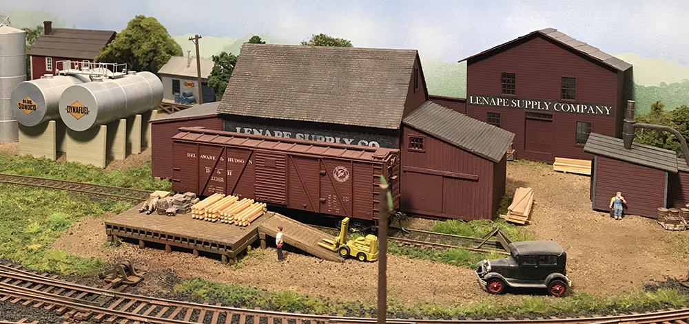 Model railroad warehouse scene showing red colored wood store houses with gray roofs and a brown boxcar along a wood loading dock.