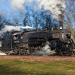 An image of the broadside of a black-painted steam locomotive trailing smoke and steam