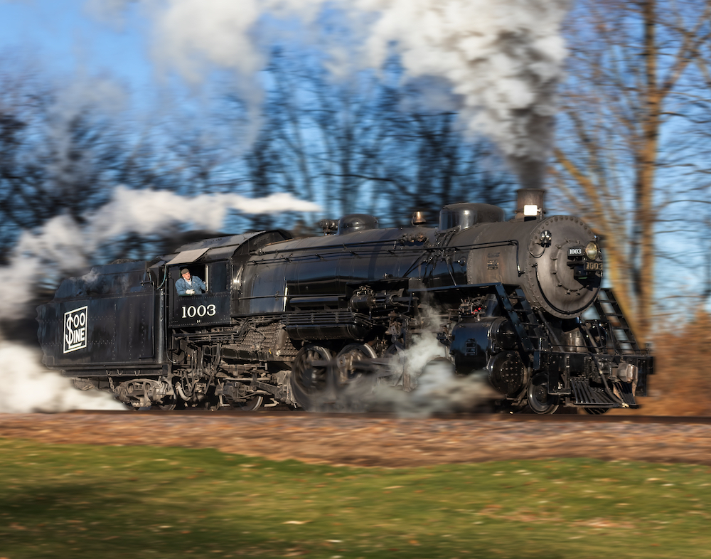 An image of the broadside of a black-painted steam locomotive trailing smoke and steam