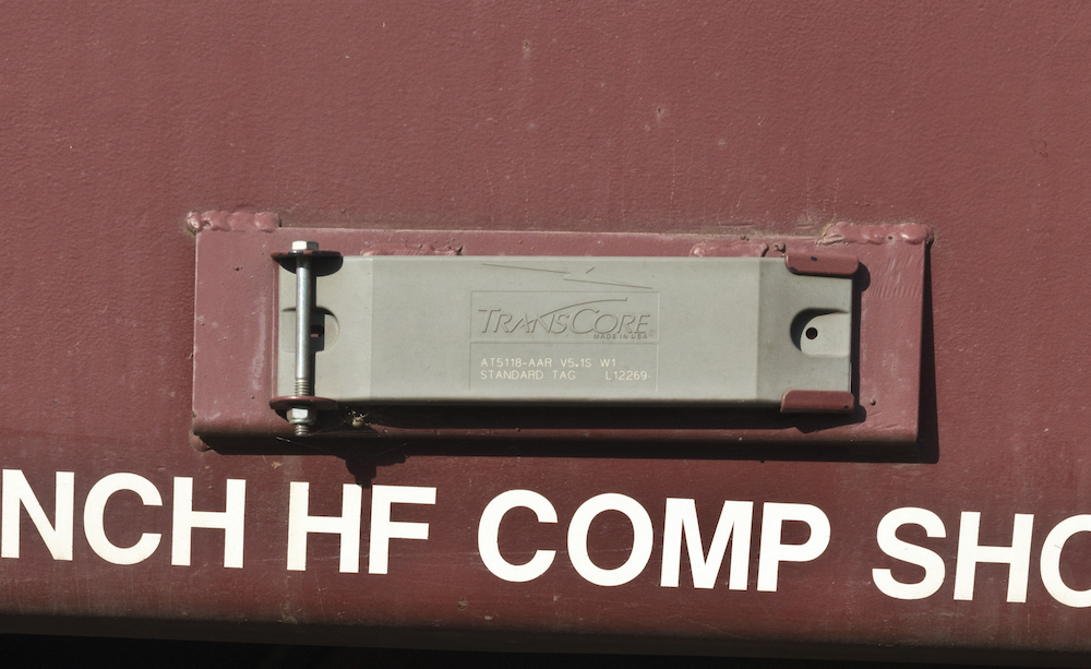 Image of a gray TransCore AEI (Automatic equipment identification) tag mounted on the side of a red railcar