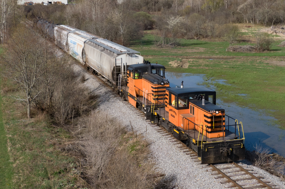 Two small, orange and black diesel locomotives roll past a flooded field