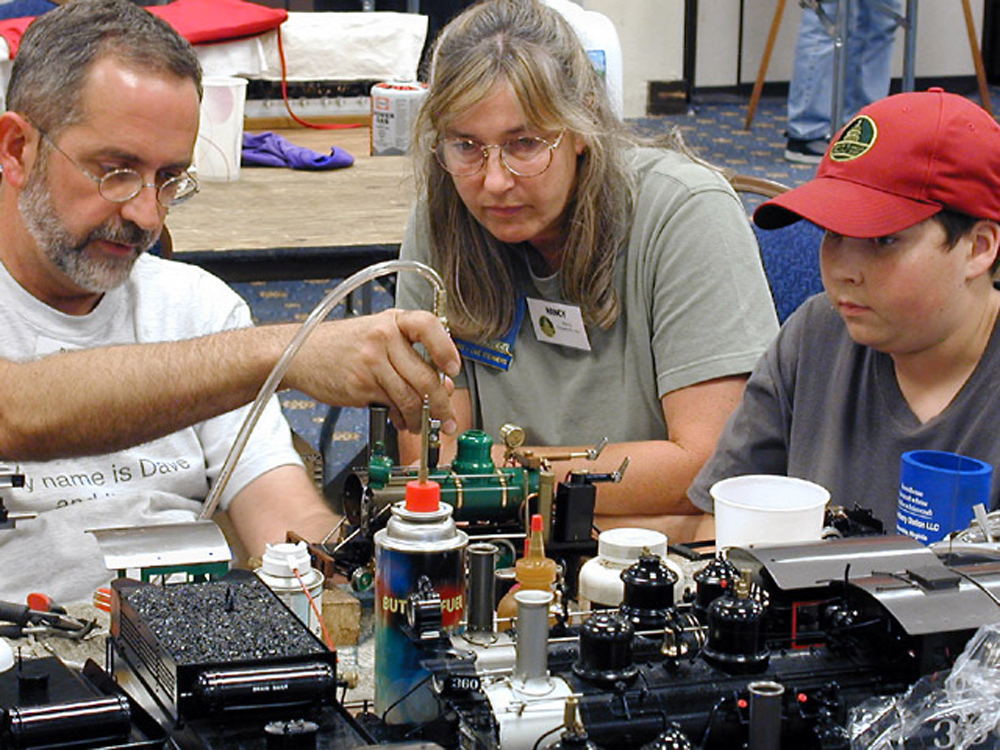 woman and boy watching man adjust live-steam locomotive model