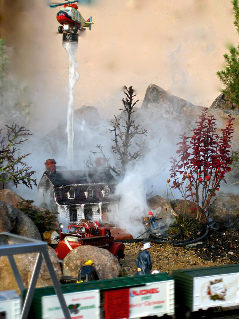 Scene of smoking house with helicopter dropping water on garden railway