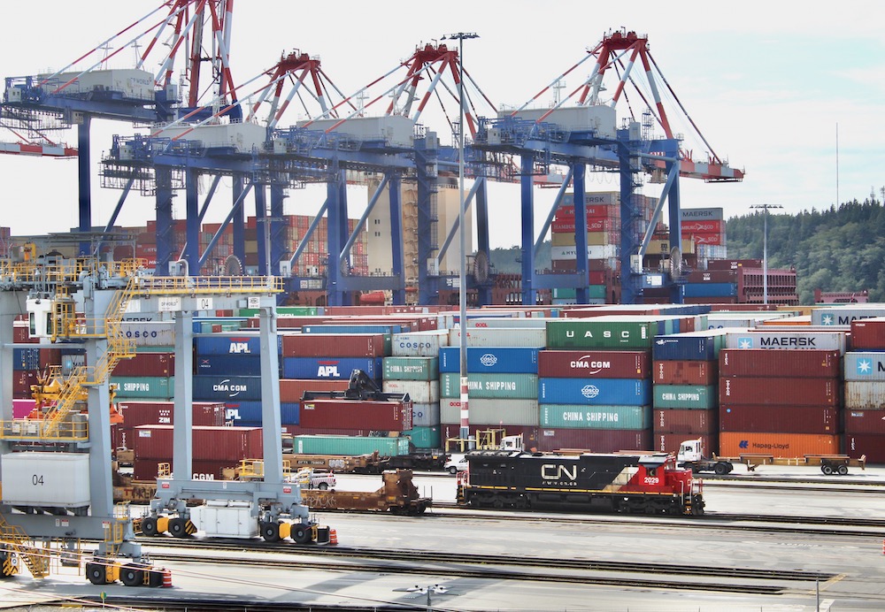 Locomotive moves intermodal cars with stacks of containers and cranes in background at port