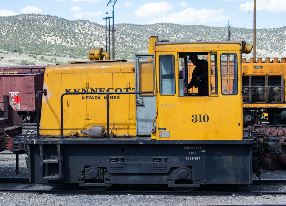 A sideview of a small yellow diesel locomotive on its side, lettered in black, are the words Kennecott Nevada Mines and the number 310