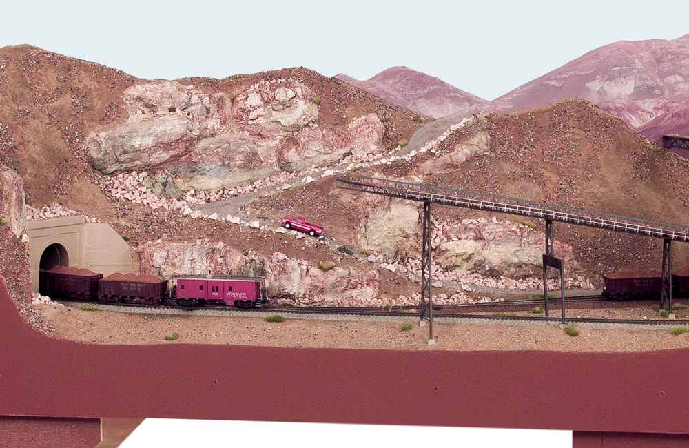 A model train rolls through a desert landscape in front of a mountain with a winding road