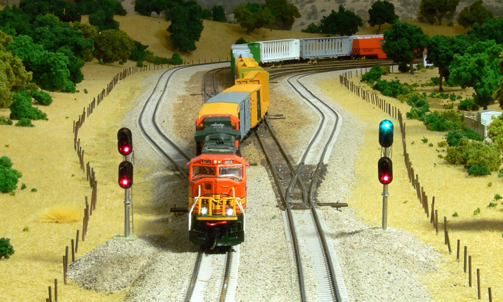 A train led by an orange diesel crosses over from one track to another between a pair of two-head signal masts