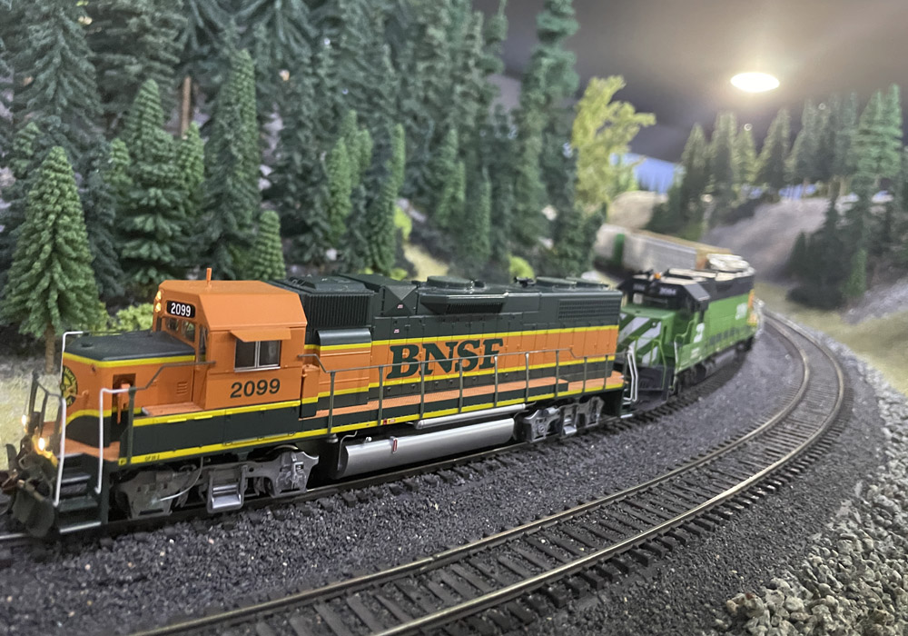 An orange-and-black BNSF diesel model leads a freight around a curve in front of a forested hill