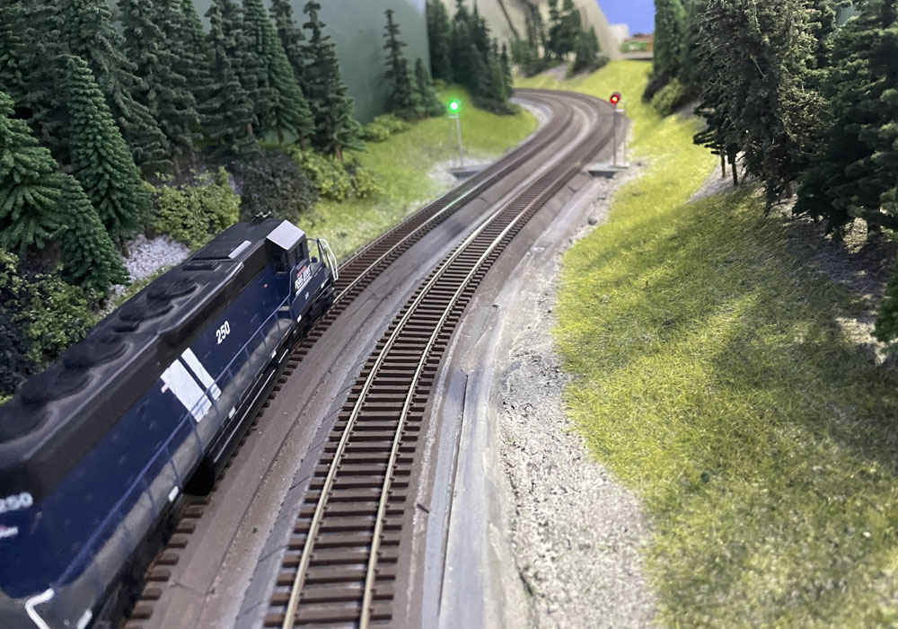 A blue HO scale locomotive approaches a green signal on a double-tracked main line in a forested setting