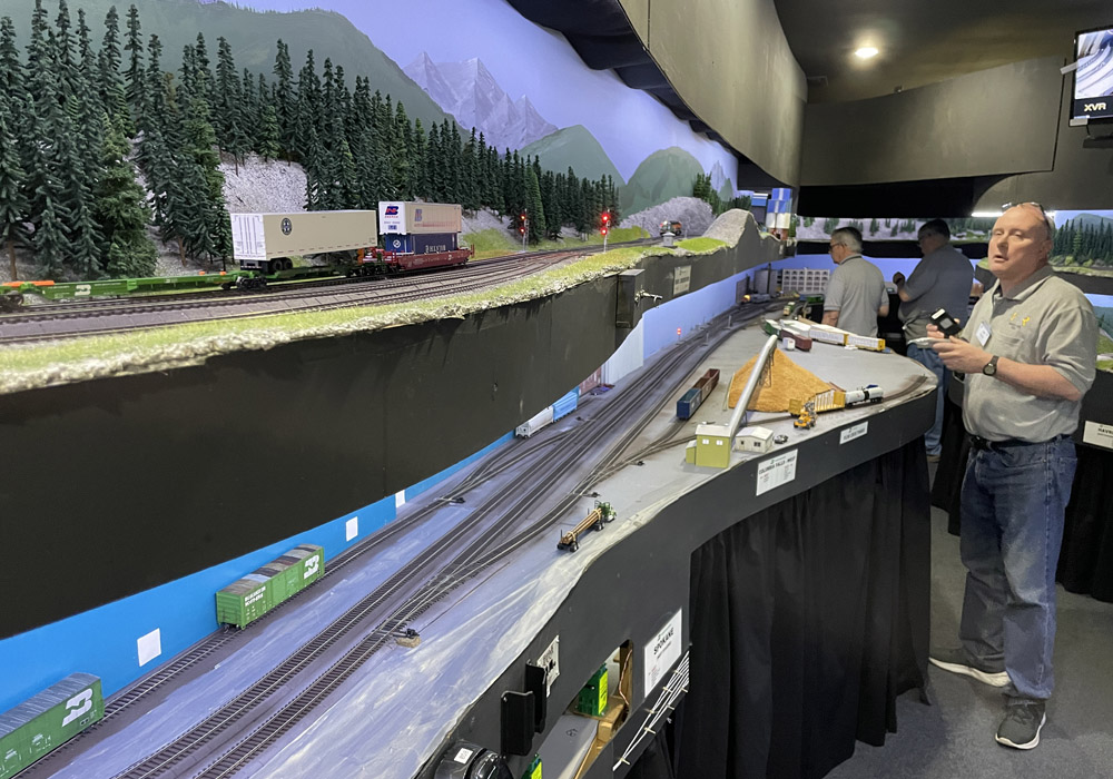 An operator in a gray shirt watches his locomotive on the upper deck of an HO scale train layout