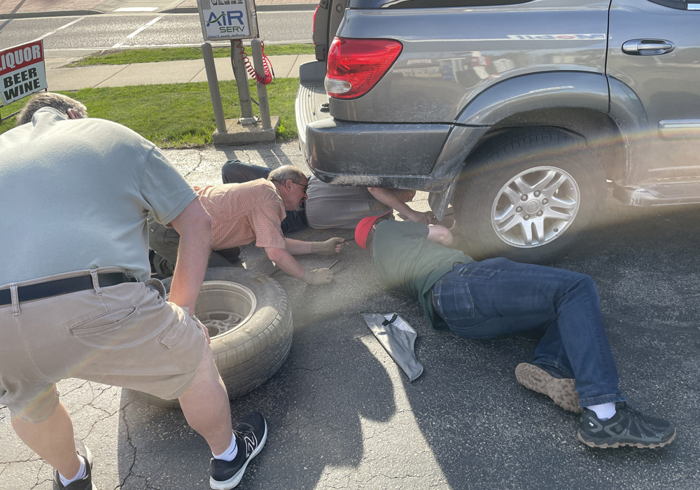 Four men work around and under the back end of a silver SUV in a parking lot