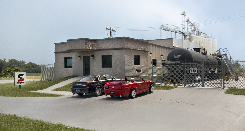 Color photo of building and storage tanks with tank cars and vehicles nearby.